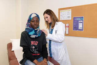 Medical Services - Image of a woman in a lab coat giving an exam to female student wearing a head scarf and NIU t-shirt